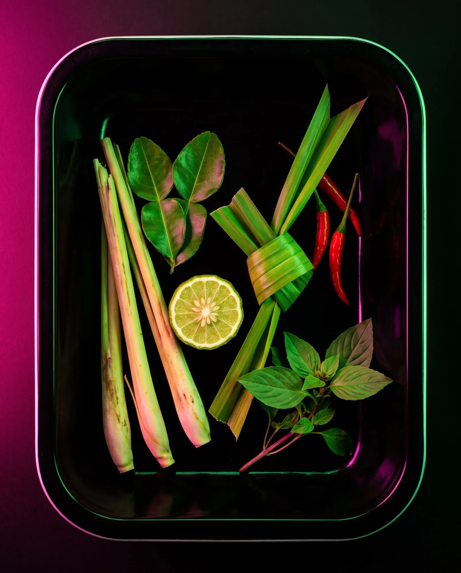 Lemongrass, makrut lime leaves, pandan and red chillies laid out on black enamel under neon