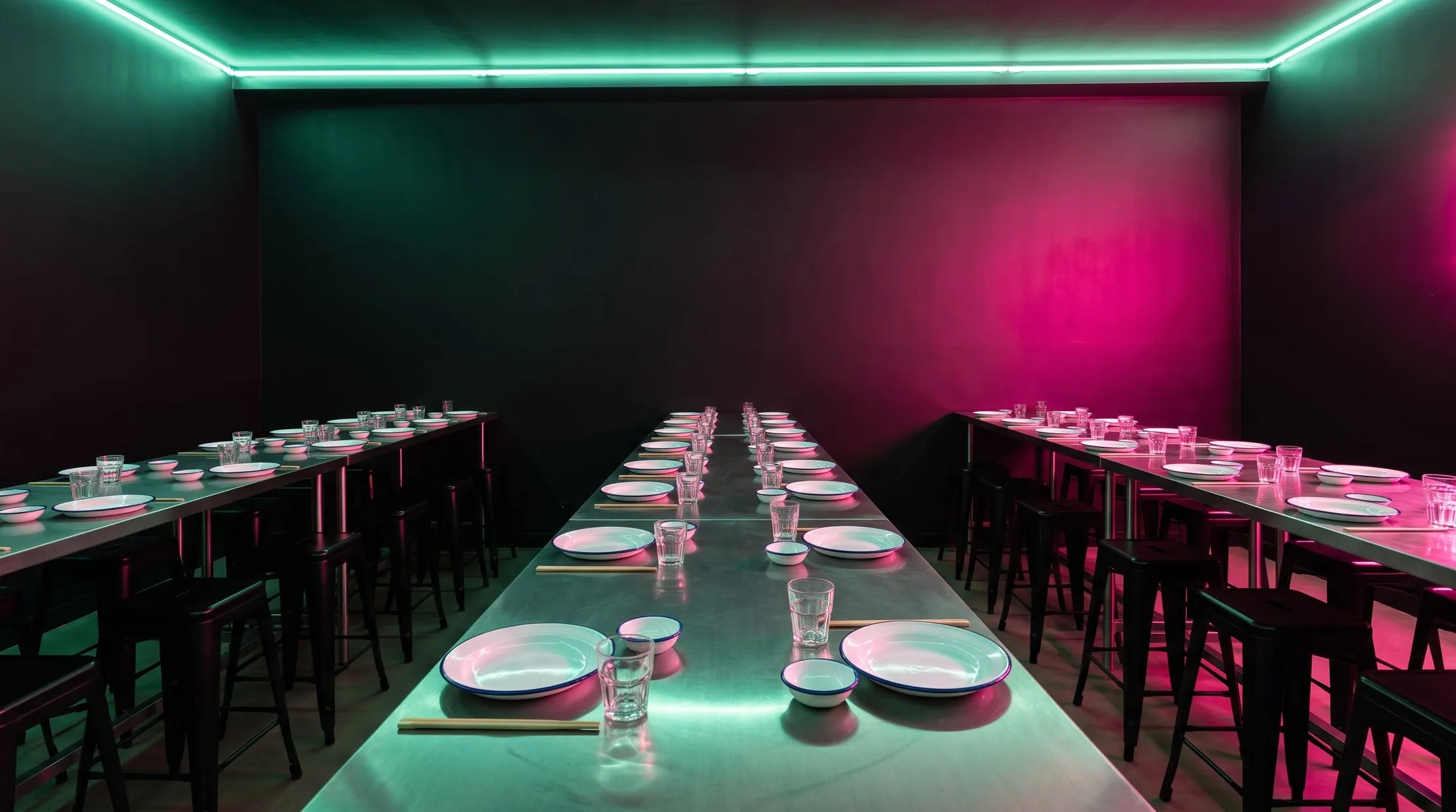 A long steel table set for a big group in a dark room under green and pink neon
