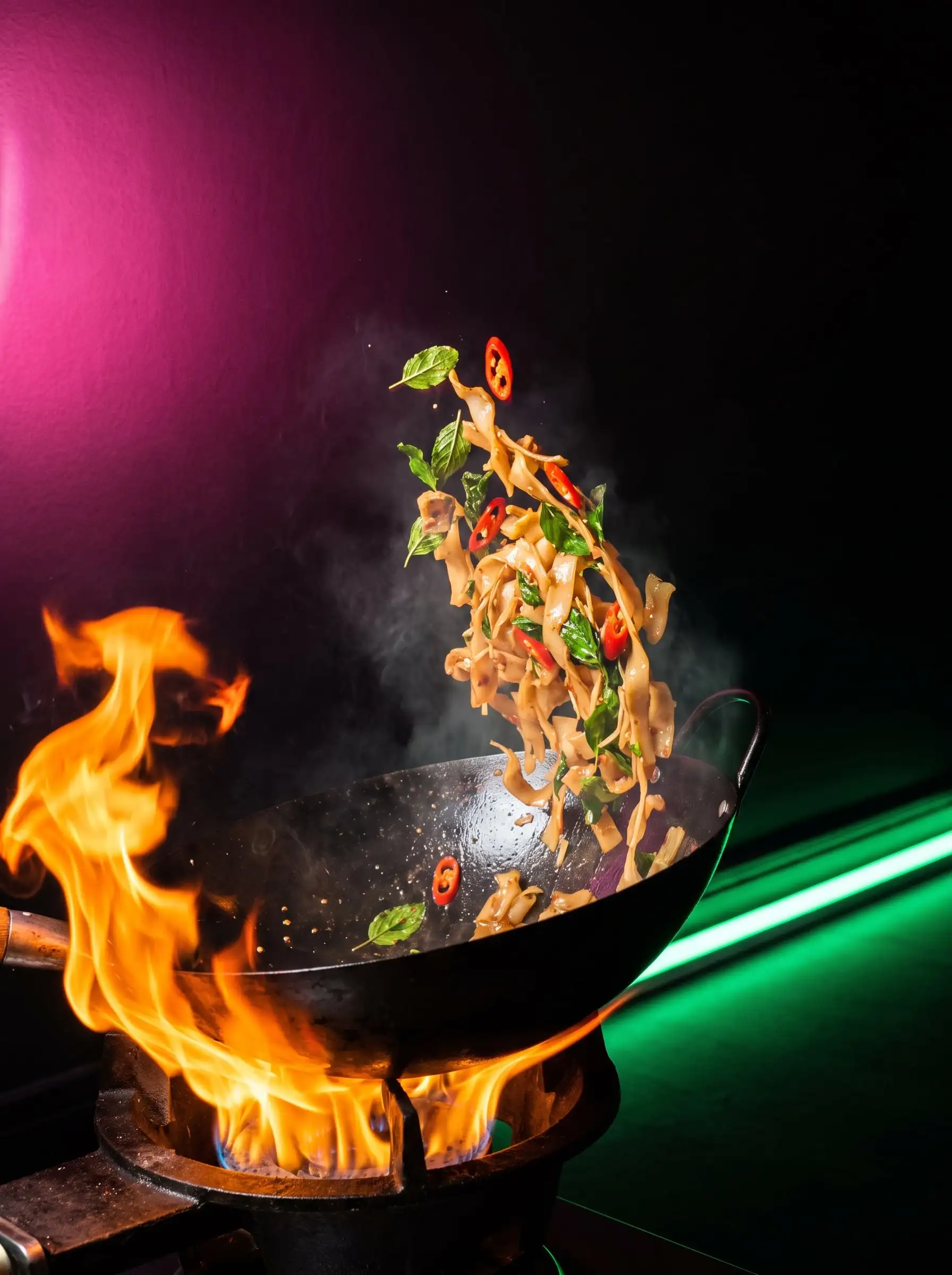 Flat rice noodles, basil and chilli flying out of a flaming wok under pink and green neon